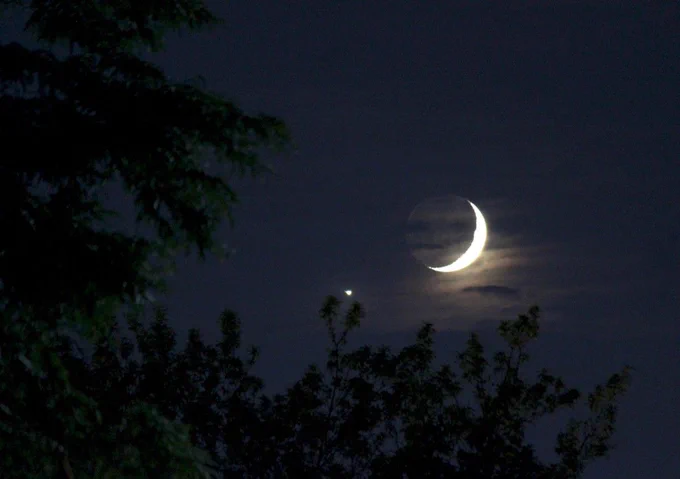 A crescent moon peeks above trees and branches in this photo of the night sky taken on July 15, 2018. Below it and to the west, the planet Venus appears as a bright dot. Credit: NASA/Bill Dunford 