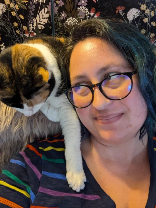 The picture is of a woman with black glasses looking over her shoulder to a cat that is sitting behind her. The cat is brown, black, and white and is reaching over the woman and their paw is on her chest.