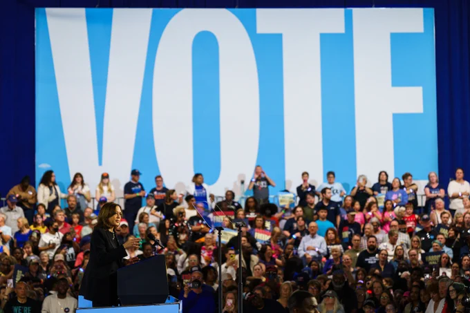 Vice President Kamala Harris delivering remarks at a campaign event. Behind her are supporters and a "VOTE" banner. 