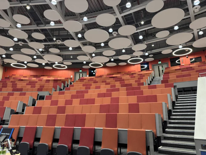 A large room. The ceiling is covered in white circles and the seats are a mix of orange and red.