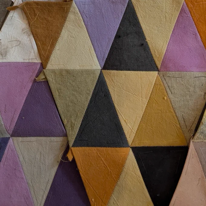 Section of patchwork fabric made up of connected isosceles triangles in shades of purple, lilac, orange, beige, dark grey