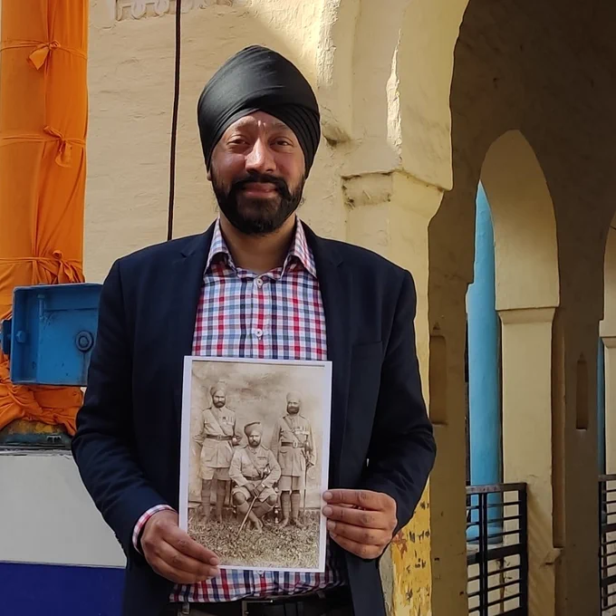 Dr Tejpal Singh Ralmill stands outside a building with yellow arches, smiling at the camera while holding a black and white photo of his great-grandfather, Subedar-Major Bawa Singh with fellow officers.