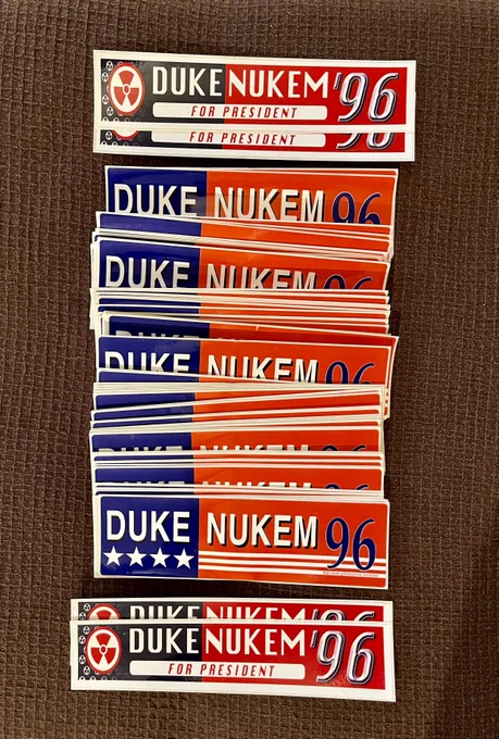 An image showcasing a collection of vintage-style bumper stickers that read “Duke Nukem ’96 For President.” The stickers are in different designs, featuring patriotic red, white, and blue colors, and some have stars or bold typography, evoking the look of a political campaign. The stickers are stacked neatly on a dark textured surface.