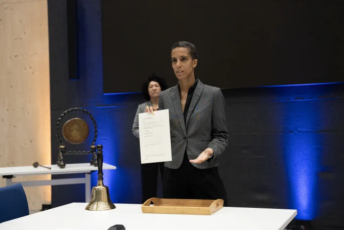 Laila and Nao are in a large room, indoors. Laila is standing at a table, holding up a contract as she speaks. A large metal bell sits on the table in front of her. Behind her, Nao stands silently by a second table with a large gong and mallet on it.