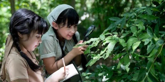 Two young girls examining the leaves on a shrub, one is looking through a magnifying glass while the other writes notes in a notebook.
