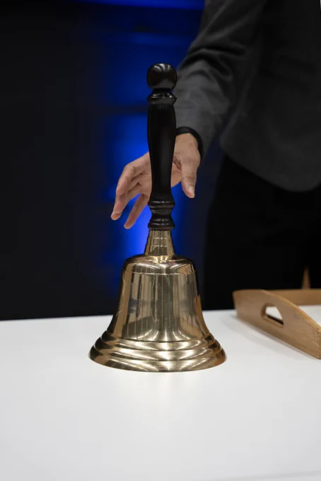 A hand reaches to grasp the handle of the bell, which sits on the white table, framed by dramatic blue lighting.
