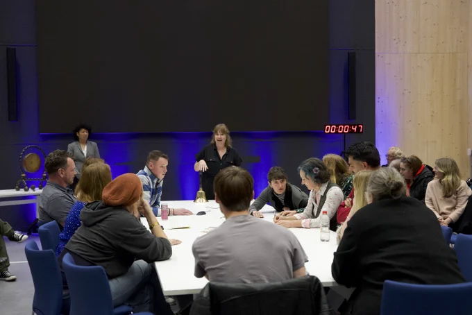 About a dozen Players sit at a long white table. One Player stands at the head, gesturing energetically. Others lean forward, speaking, or holding their hands to their mouths. The clock marks 47 seconds left.