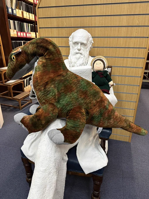 A large cuddly dinosaur sits on the knee of a seated life sized model of Charles Darwin. Whilst a knitted Mary Anning sits on his arm.