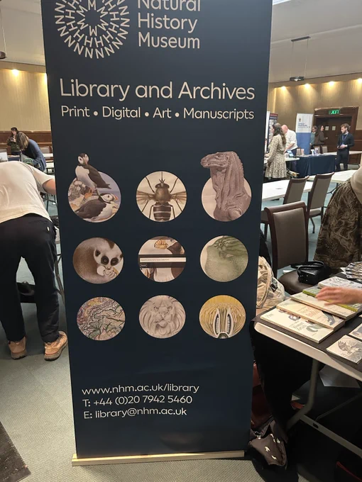 Large, black,  free standing banner headed 'Library and archives', stands in a room with people setting up tables and chairs.