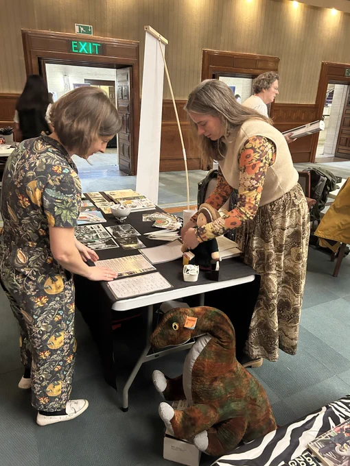 Two people organising a table with books and handouts displayed on it. On the floor next to the table is a large dinosaur cuddly toy.