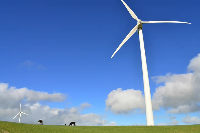 A blue cloudy sky and stretch of green field with a wind turbine in the foreground and one in the background. Three cows graze in between them in the distance.