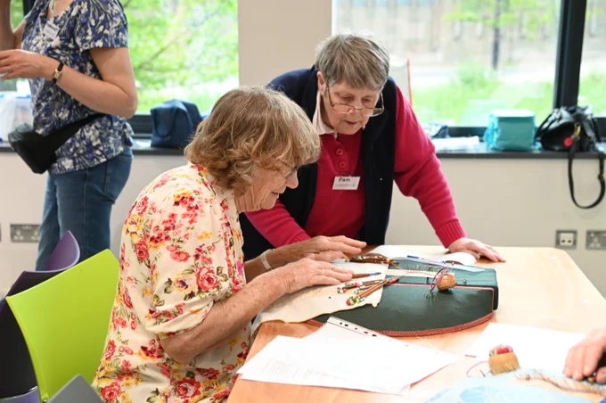 A woman sitting in front of a lace-making cushion with another woman watching.