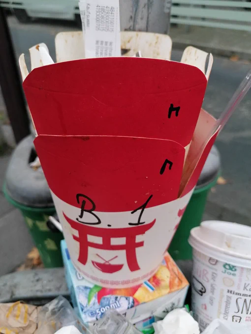 Foto von einem Mülleimer, in dem eine rot weiße Fastfood Verpackung liegt, darauf ein torii und eine Schale 