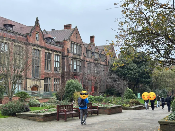 Old red brick uni campus with trees. People are walking on a paved path.