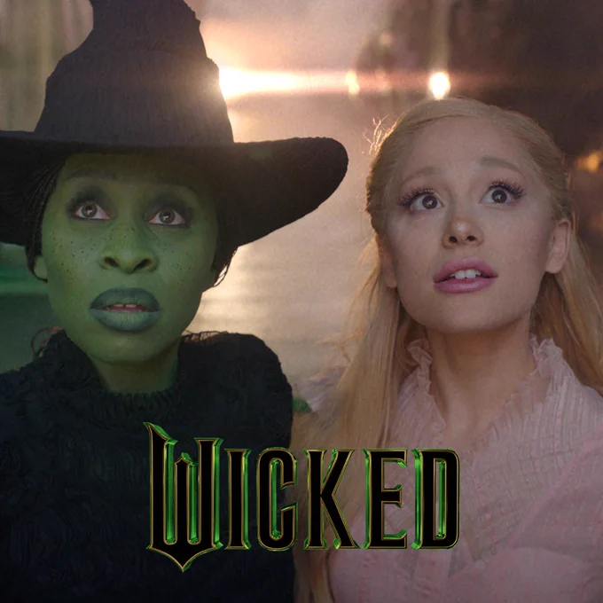 An image from the film Wicked, featuring two women in mid shot, looking upwards. The woman on the left is a witch, wearing a black dress and black hat, and her skin is green. On the right the woman is wearing a pink dress and has long blonde hair. The graphic title Wicked is placed at the bottom centre frame, and is written in black capital letters, highlighted in illuminous green. There is a light shining in the background.