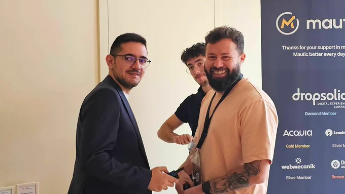 Two men are shaking hands, smiling at the camera. One man is wearing a dark suit and glasses, while the other is in a beige shirt with visible tattoos. In the background, a third person is smiling near a promotional banner featuring various sponsors.