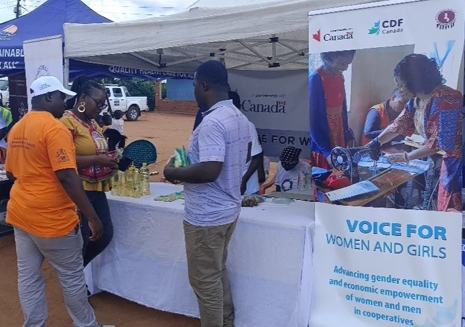 Two men and a women standing in front of a table looking at items. To their right is a VOICE for Women and Girls pull-down banner.