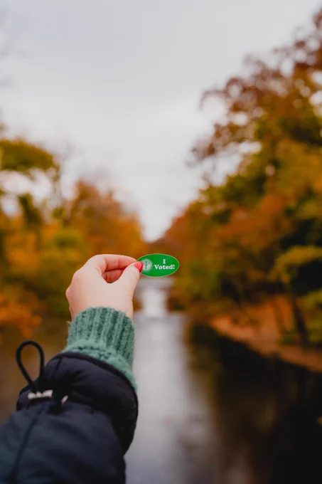 A person's hand is holding a green sticker that reads, "I voted," in front of the Red Cedar on a fall day.