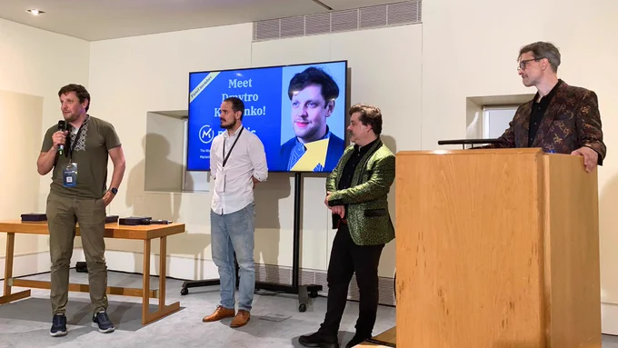 Four speakers are on stage at an event. One man is presenting with a microphone, while the others stand beside him. A screen in the background displays a portrait and text. The setting appears to be a conference room, with a podium in the foreground.