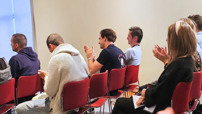 A group of people seated in red chairs, clapping and reacting to a presentation. The focus is on the audience, with some individuals visible from the back and side, wearing casual clothing. The setting appears to be an indoor space with neutral-colored walls.