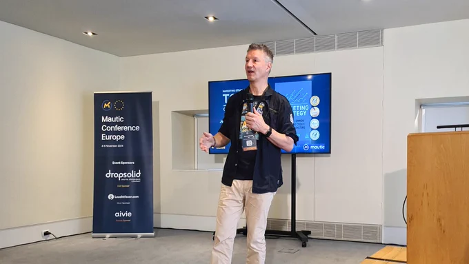 A speaker stands on stage, presenting at the Mautic Conference Europe. In the background, a digital screen displays conference-related graphics. A banner with logos for various sponsors is positioned to the left.