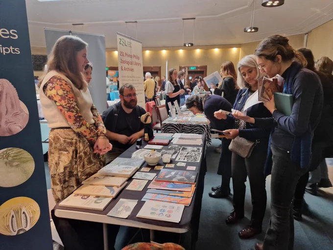 A busy room of people sitting behind tables advertising their institution. People stand in front of the tables looking at what is displayed and chatting to those at the table.