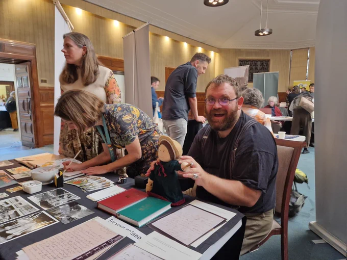 Three people behind a table, with books and paper items on it. Two of the people are standing and one is seated looking at the camera holding a knitted Mary Anning doll.