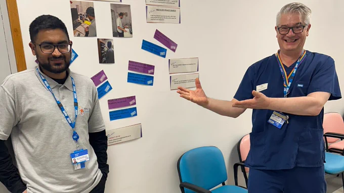 Daniyal smiling with a male charge nurse in a blue uniform, stood outside the department with photos and information about Project Search displayed on the wall
