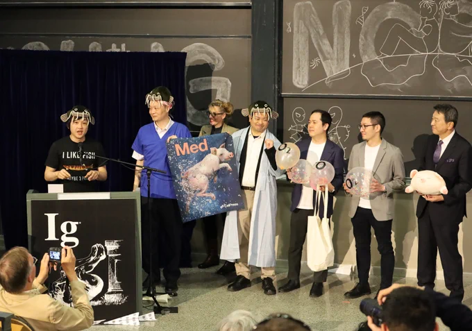 Takanori Takebe and his research team stand on stage wearing anime sea creature hats and holding balloons with O2 written on them and the Med cover for the issue their award-winning research was published in, which features a pig underwater with bubbles floating up from its butt.