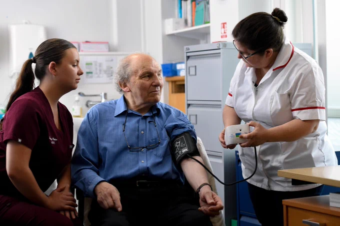 Two healthcare professionals are assisting an elderly patient with a blood pressure check in a clinic setting.