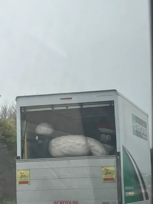 A truck on the road transporting large sculptures of Shaun the Sheep visible through the rear window.