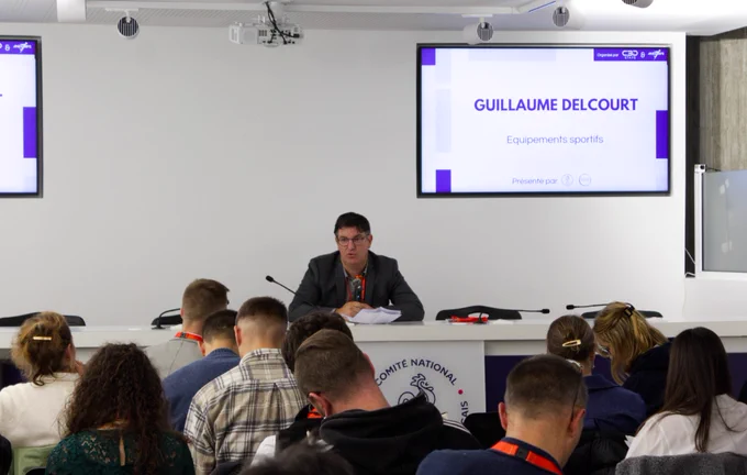 Le chercheur est assis au centre de la table de conférence avec écrit sur l'écran derrière lui, "Guillaume Delcourt, Equipement sportifs"