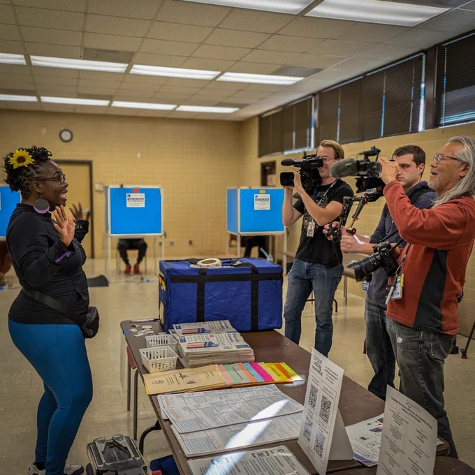 The image shows Dr. Flo Cofer speaking to a group of journalists and photographers in a voting area. She stands near a table filled with voting materials, ballots, and information sheets. Dr. Cofer is wearing a black top, blue leggings, and has a yellow flower in her hair. She gestures expressively with her hands while talking. Three media personnel stand in front of her, two holding cameras and one with a boom microphone, capturing the moment. In the background, there are blue voting booths with people seated at them. The setting is indoors, with beige-tiled walls and fluorescent ceiling lights.