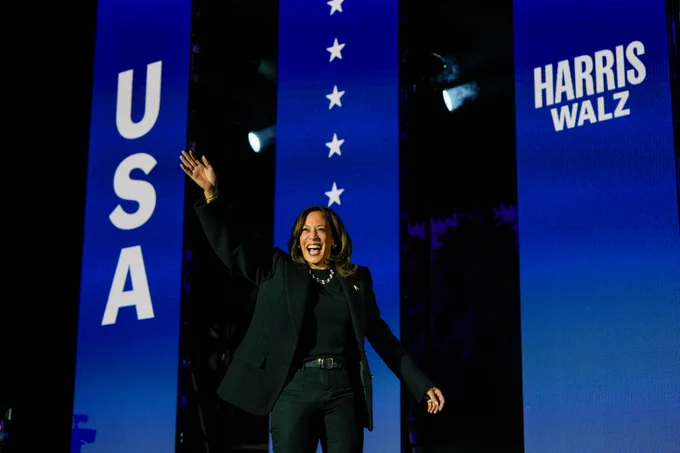 Vice President Kamala Harris waves enthusiastically on stage at a rally, standing between signs that read "USA" and "Harris Walz.