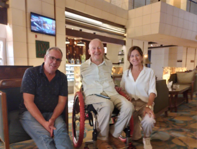 Marie is kneeling next to David Constantine, ISPO President who is seated in a wheelchair next to Claude Tardif, ISPO's Past President.  They are sitting in a hotel lobby.
