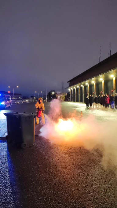 Palokuntanuorten toimintanäytöksessä nuori sammuttamassa "roskalaatikkopaloa" käsisammuttimella. Kurssilaiset seuraavat.