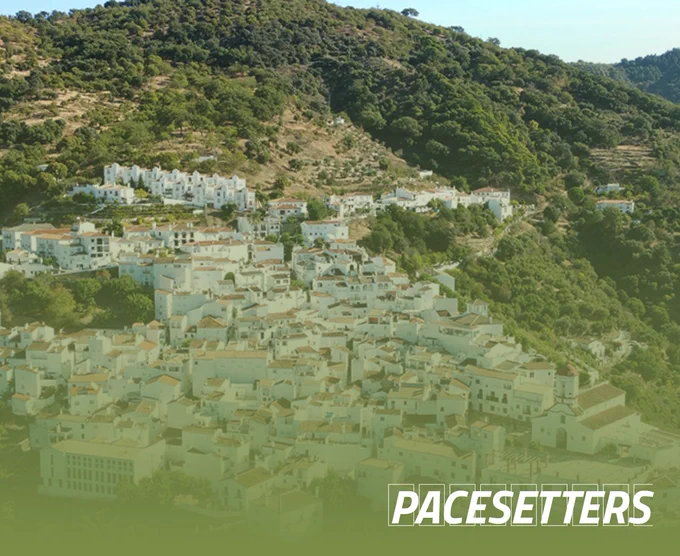 A village on a mountain side. Nearly every building is white with a clay colored roof. It looks small but vibrant, is very tightly built and bucolic. Around it a lot of green trees. The image has a green gradient flowing from the bottom, with the pacesetters logo in the bottom right corner.