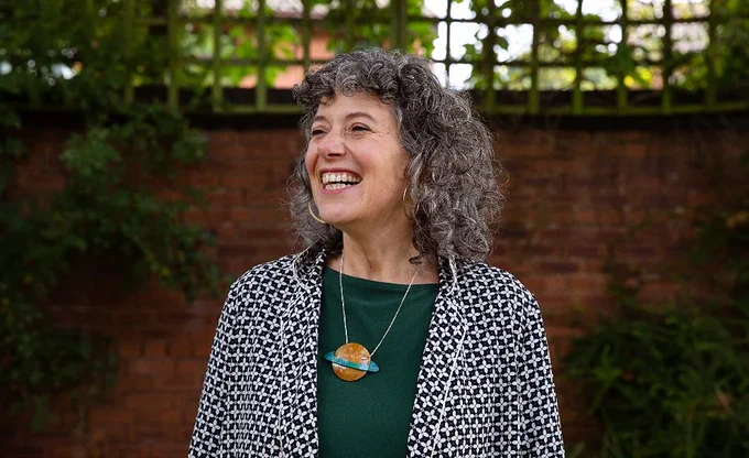 Julia Samuels is a white woman, with grey curly shoulder length hair. She wears a dark green tshirts, a blazer with a black and white check, and a necklace with a large planet pendant. She is laughing and looking to her right. Behind Julia is a brick wall with a wooden trellis on top, and various plants climbing it.