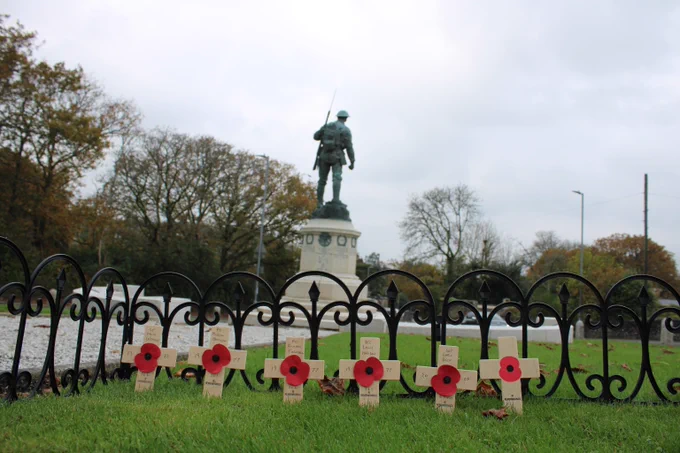 In the foreground are six small wooden crosses with a poppy at the centre of each. The crosses are surrounded by a small black fence. In the background is a war memorial depicting a soldier with a rifle over his shoulder.