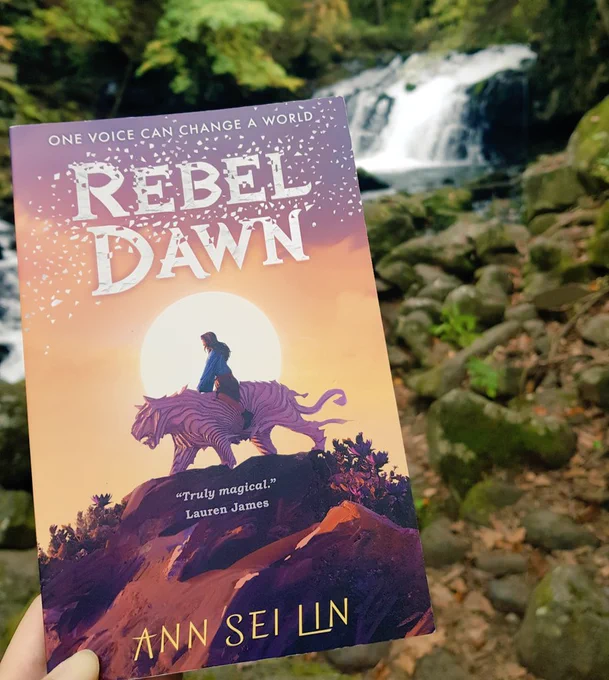 Rebel Dawn held up against a backdrop with a waterfall in the middle of a forest 