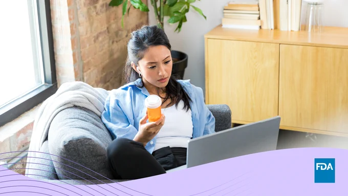 A woman of Asian American descent holds a bottle of medicine as she looks at a computer screen. 