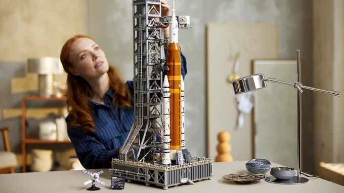 A woman builds the NASA SLS rocket which is shown in the forefront. The rocket is orange with white boosters and stands on a grey model of the launch pad.