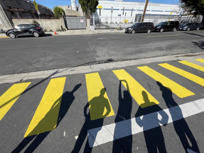 The shadows of five crosswalk painters shown in front of a freshly painted yellow crosswalk. 
