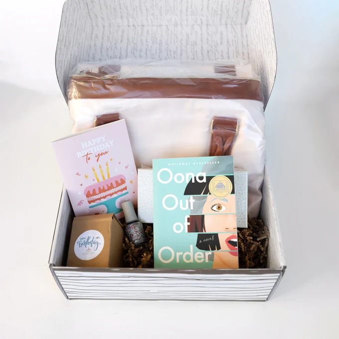 An open box with a book, greeting card, nail polish, a box, and a folded bag