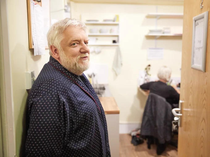Simon Russell Beale wears a blue dressing gown and stands in a doorway of a dressing room.