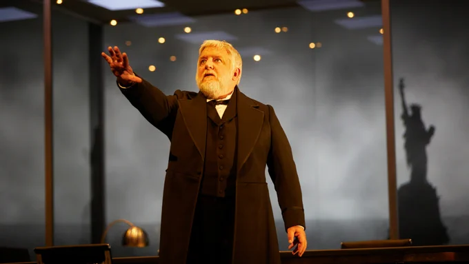 Simon Russell Beale wears a black suit and stands in an office room in front of the statue of liberty.
