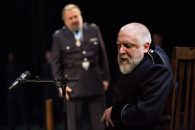 Simon Russell Beale wears a military jacket and sits on a chair next to a microphone. A man stands behind him.