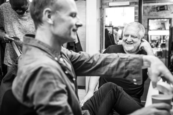 Simon Russell Beale sits on a chair and laughs with another man who sits next to him in a dressing room.