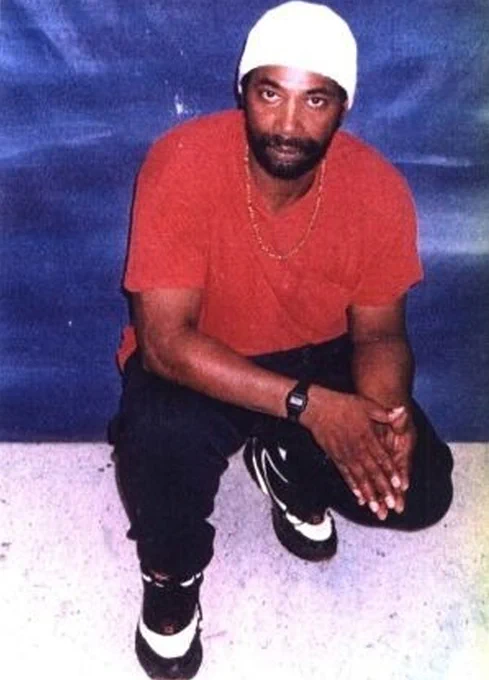 A New Afrikan man crouches down. He is wearing a red t-shirt, dark trousers and a white beanie.