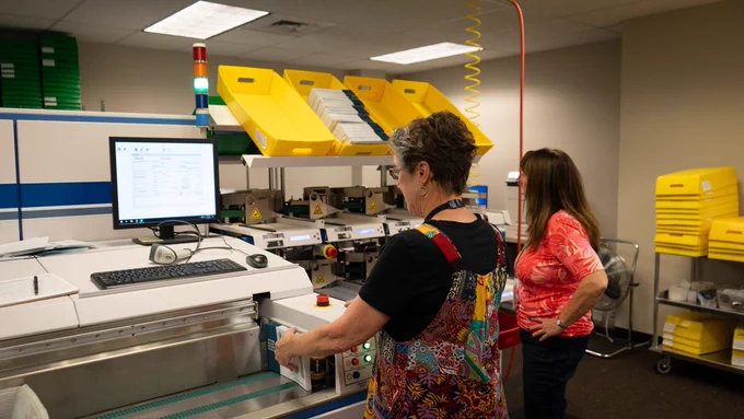 Two election workers run ballots through the sorting machine for signature verification, marking ballots as returned and grouping them into batches for further processing.